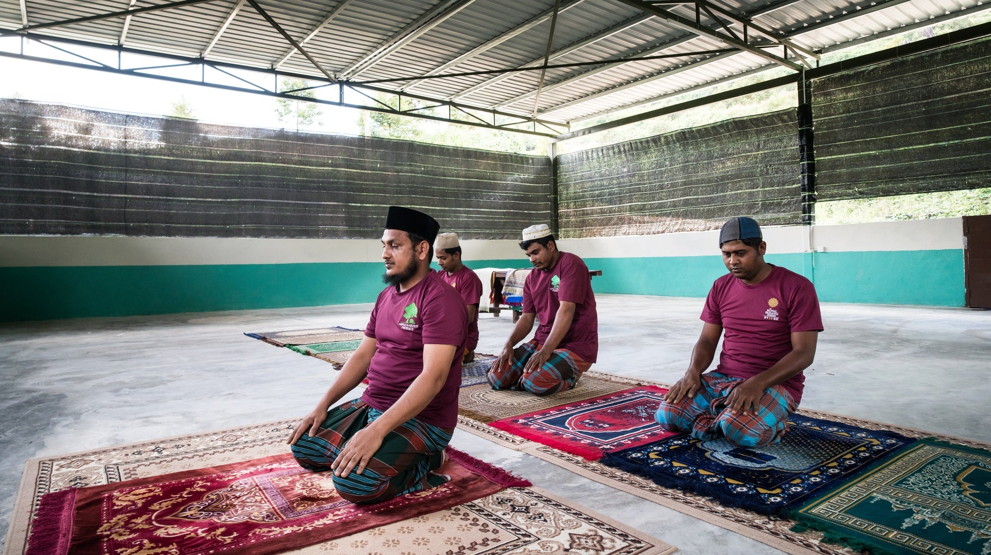 Four men in maroon shirts and caps sitting on prayer mats in a mosque.